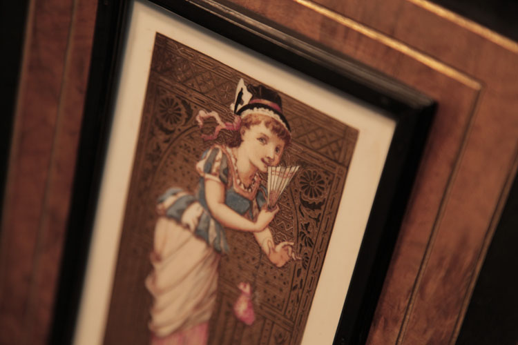 Detail of the hand-painted ceramic tile mounted on piano cabinet edged with a black border and gold embossed, patterned background. Tile depicts a coy girl holding a fan and purse 
