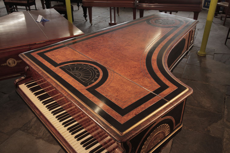 Piano lid is in burr walnut with black dual border and carved decoration in low relief. At the piano tail, a circular motif bordered with spiralling rope motif contains four symmetrical anthemions. Above the piano fall, a semi-circular sun motif is bordered with stylised swags and lotus leaves