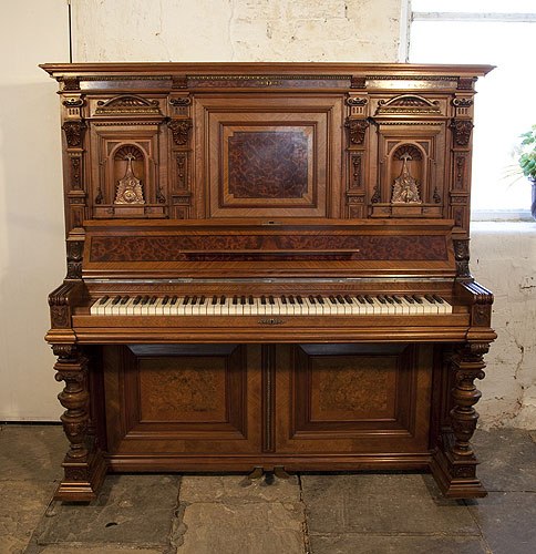 Golden Age of Pianos. A German upright piano with a Neoclassical style walnut case and cup and cover legs. Cabinet features ornately carved pilasters in high reief and copper sconces in a sea monster design